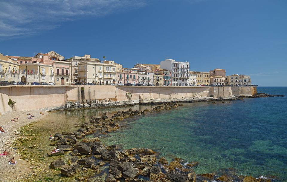 Case e mura del lungomare di Ortigia sopra una piccola riva di ciottoli e scogli bagnati dal mare